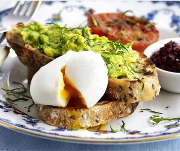 Plate with smashed avocado on toast, poached egg, grilled tomato, and a small dish of relish, garnished with herbs.