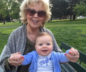 Elderly woman with sunglasses and short hair holds a smiling baby in a blue sweater on a park bench, surrounded by greenery.