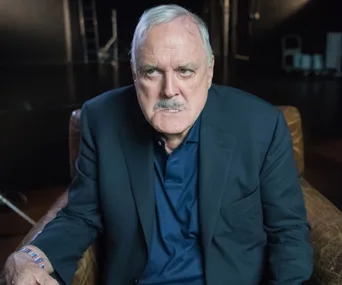 Man with a serious expression, wearing a dark suit, seated in a dimly lit room.