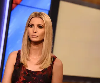 A woman with long blonde hair in a sleeveless red and black dress, sitting in a studio with blurred background.