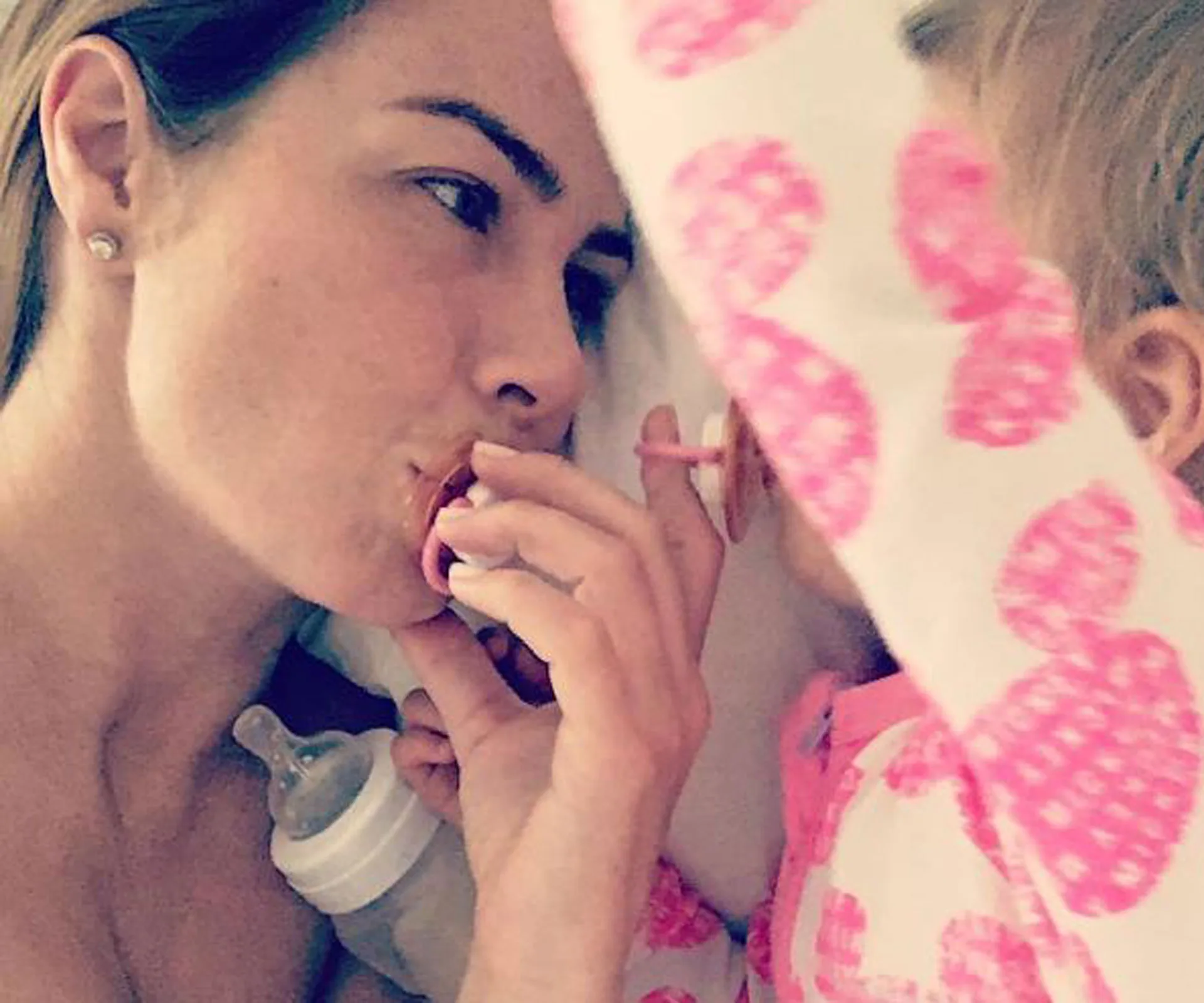 Mother and child cuddle in bed; mother holds a pacifier with a milk bottle nearby.