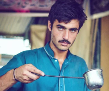 A man in a blue shirt pours tea from a metal cup, with an intense expression.