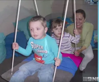 Children on a swing with an adult smiling in the background in a cozy room setting.