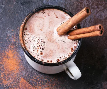 Mug of hot chocolate topped with cinnamon sticks, on a dark textured surface with cinnamon powder scattered around.