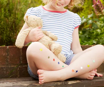 Child in striped shirt holding a teddy bear, with colorful star stickers on their legs, sitting outside on a wooden bench.