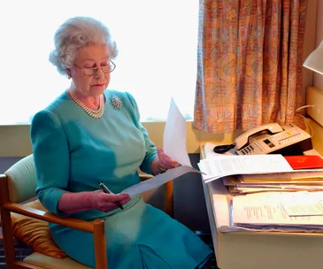 Elderly woman in blue dress seated, reviewing papers at a desk with a telephone and documents.