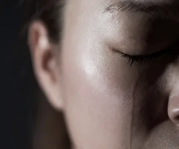 Close-up of a person's face with eyes closed, tear rolling down cheek against a dark background.