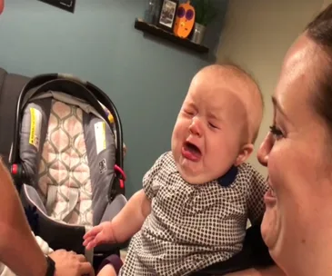 Baby cries while being held, reacting to parents' interaction; car seat visible in background.