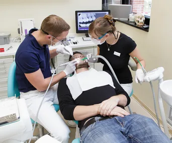 "Dentist and assistant treating a patient in a dental office with dental tools and X-ray on screen."