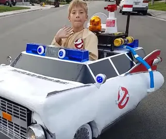 Child in a Ghostbusters costume sits in a wheelchair transformed into the Ecto-1 car on a neighborhood street.