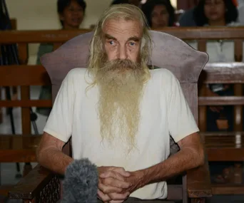 An elderly man with long white hair and beard sits in a courtroom, hands clasped, with people in the background.