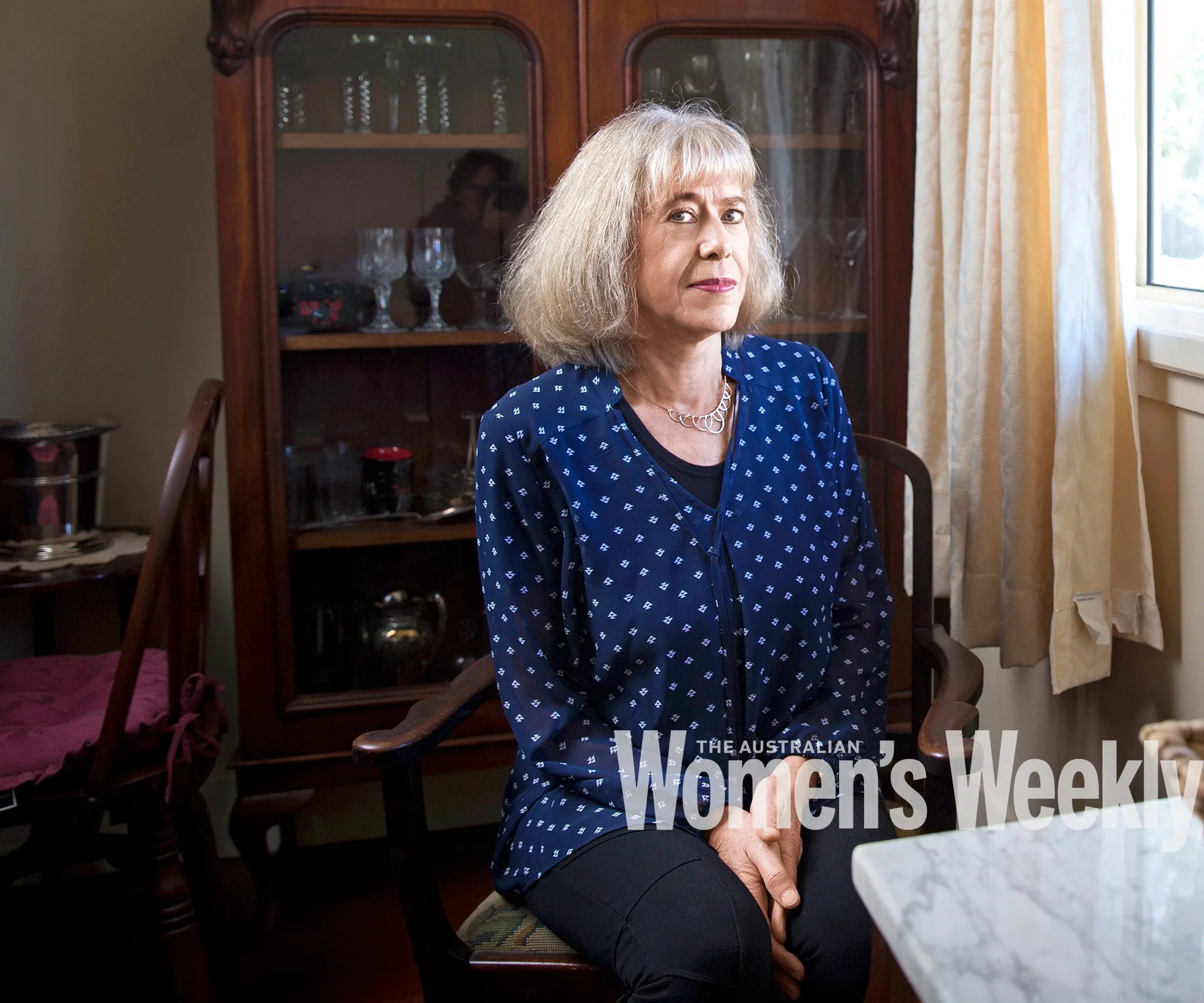 A woman with short hair in a blue polka dot blouse sits in a dining room with glass cabinet behind, sunlight through window.