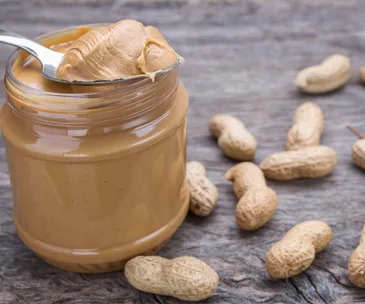 Jar of peanut butter with a spoonful beside scattered peanuts on a wooden surface.
