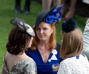 "Three women in formal attire conversing at an outdoor event, focusing on the woman in a blue dress with a decorative hat."