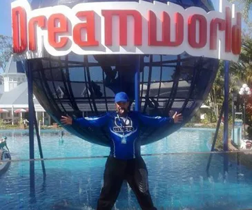 Man in a blue shirt poses playfully in front of a Dreamworld sign by a water feature.