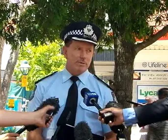 Police officer giving a press conference, surrounded by microphones, with trees and signs in the background.