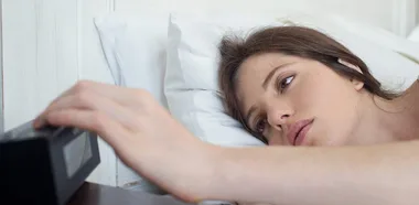 A tired woman lies in bed, reaching for an alarm clock.