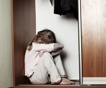 A young girl sitting in a closet with her head resting on her knees, wearing a pink top and light pants, looking distressed.