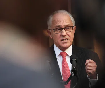 A person speaking at a podium, wearing glasses and a suit with a red tie, in an outdoor setting.