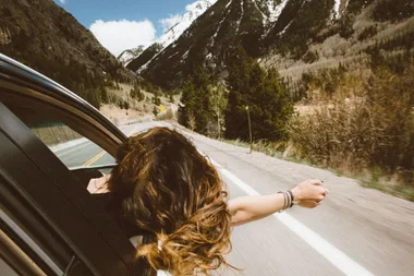 Person with long hair leans out of a car window, enjoying a scenic mountain road.