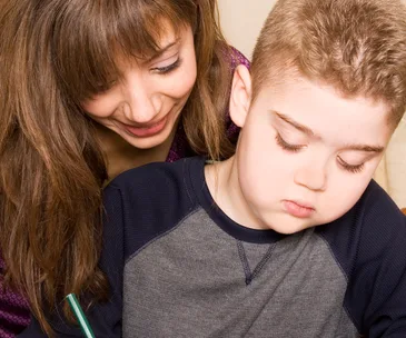 A woman smiles while assisting a young boy with a task, the boy focuses intently, holding a pencil.