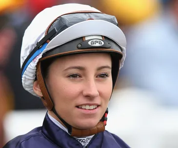 Female jockey wearing a helmet and racing attire, smiling, at a horse racing event.