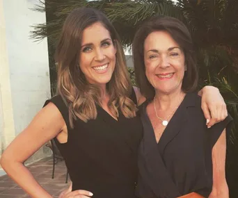 Two women smiling and embracing outdoors, with palm leaves in the background.
