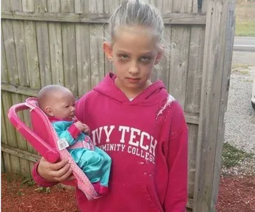 Young girl dressed as a tired mom for Halloween, holding a baby doll in a car seat, wearing a pink Ivy Tech hoodie.