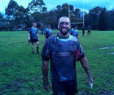 Rugby player smiling, covered in mud, standing on a lush field with teammates and goalposts in the background.