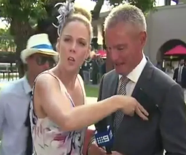 Racegoer interrupts live TV at Melbourne Cup to return lost wallet, pointing towards a suited man.