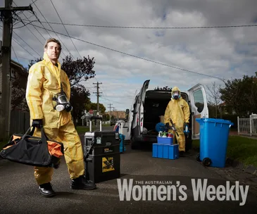 Two people in yellow hazmat suits clean near a van on a suburban street, with equipment and blue bins visible.