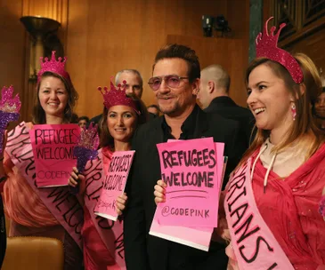 Bono poses with activists wearing pink, holding "Refugees Welcome" signs at an event.