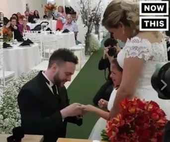 Groom kneels to give a ring to his teary-eyed stepdaughter at a wedding ceremony, with bride watching beside them.