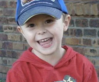 A young child in a red hoodie and blue cap, smiling in front of a brick wall.