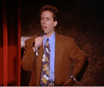 Comedian performing on stage, wearing a brown jacket and patterned tie, holding a microphone.
