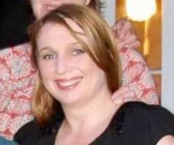 A woman with brown hair smiling in a close-up, wearing a black top, with a person standing behind her.