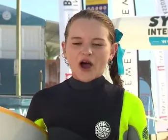 A young girl in a wetsuit, with eyes closed, likely making a humorous expression during an outdoor interview.
