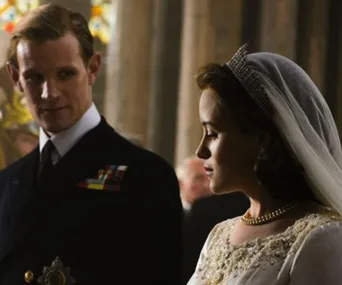 A bride stands beside a man in uniform inside a dimly lit building with colorful stained glass in the background.