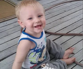 Toddler smiling while sitting on a wooden deck, wearing a tank top and pants.