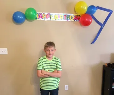 Young boy in a striped shirt stands under a "Happy Birthday" banner with balloons on a wall.