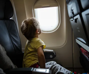 Young child in yellow shirt looking out airplane window, seated in economy class.
