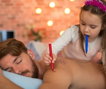Father sleeping while young daughter doodles on his back with markers.