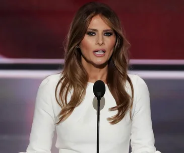 A woman in a white dress speaks at a podium with a microphone, delivering a speech.