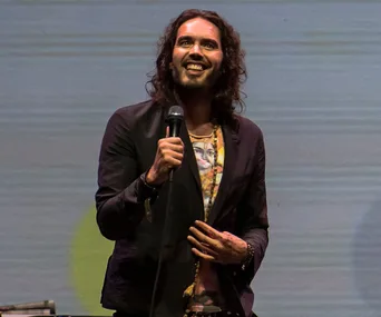 Comedian wearing a graphic shirt and blazer, holding a microphone, smiling on stage against a simple backdrop.