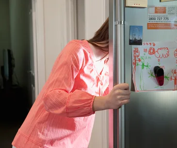 A woman in a pink top looks into a fridge with children's drawings and emergency contact info on the door.