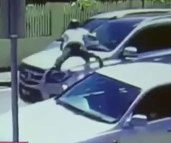A person clings to the hood of a moving SUV, surrounded by other cars on a street.