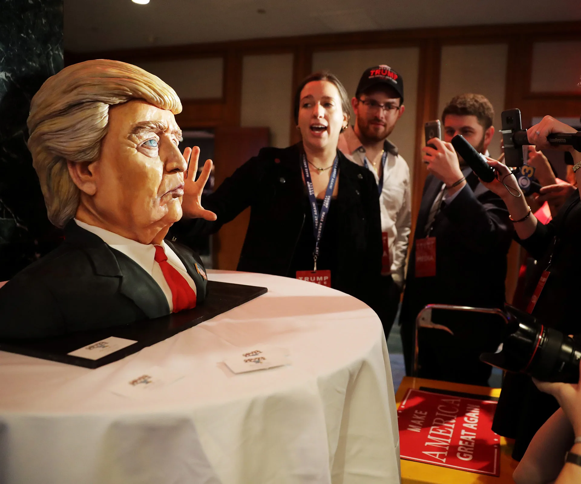 Donald Trump bust cake at event, people around taking photos.