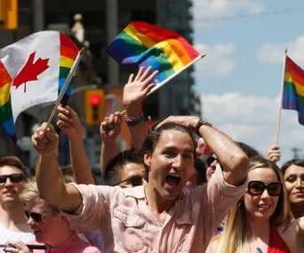 Crowd celebrating at a parade, waving rainbow and Canada flags, with a sunny sky in the background.