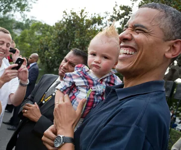 Former President Barack Obama smiling while holding a baby with a mohawk, surrounded by people taking photos.