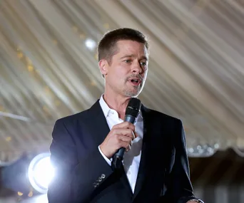 A person in a suit speaks into a microphone at an event with a well-lit backdrop and overhead drapery.